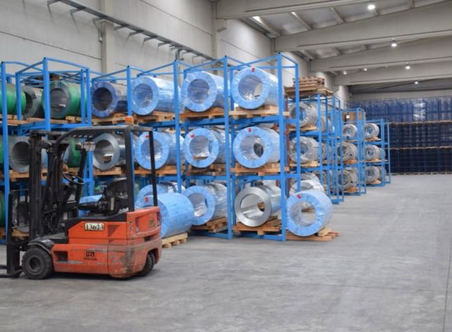 Interior de un almacén industrial logístico en Castilla-La Mancha con estanterías azules llenas de bobinas de acero envuelvas y una carretilla elevadora en primer plano.