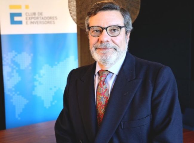 Retrato en primer plano de Antonio Bonet, presidente del Club de Exportadores e Inversores, vistiendo traje oscuro, gafas y corbata roja con motivos estampados, frente a un tótem corporativo con un mapa del mundo difuminado.