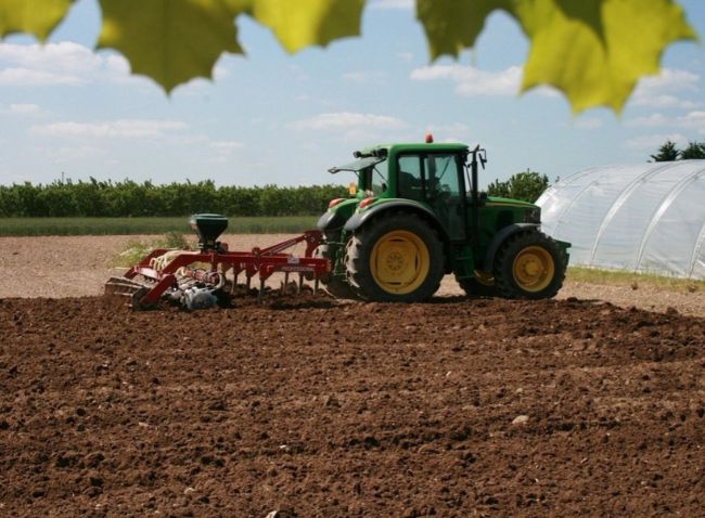 Maquinaria agrícola aplicando abono en un extenso campo de cultivo europeo, representando el impacto del precio de los fertilizantes en la producción de alimentos.