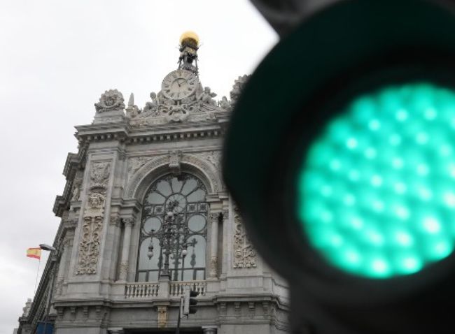Fachada clásica del edificio del Banco de España en Madrid vista por detrás de un semáforo con la luz verde encendida y desenfocada, bajo un cielo nublado.